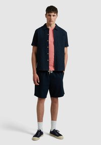 Kurzärmliges, strukturiertes, marineblaues Hemd mit Holzknöpfen, darunter ein korallenfarbenes T-Shirt, marineblaue Shorts, weiße Socken und dunkle Sneakers.