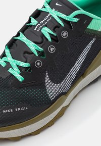 Zwarte en groene trailrunner met een gestructureerde mesh bovenkant, reflecterende accenten, brede veters en een robuuste zool. Kenmerkt zich door het Nike-logo aan de zijkant.