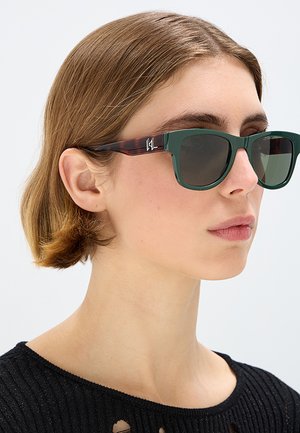 Giovane con capelli castani corti indossa occhiali da sole verde scuro con aste marrone tartarugate e una maglietta nera a maglia.