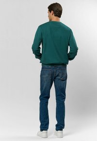 Zaļš džemperis ar apaļu kaklu un garām piedurknēm, kombinēts ar ziliem džinsiem. Redzamas baltas kedas ar gludu tekstūru un klasisku dizainu.