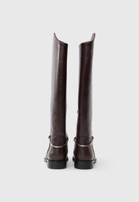 Ζευγάρι ψηλές σκούρες καφέ δερμάτινες μπότες με ασημένιους μεταλλικούς κρίκους γύρω από τις φτέρνες, εμφανιζόμενες από πίσω σε απλό λευκό φόντο.