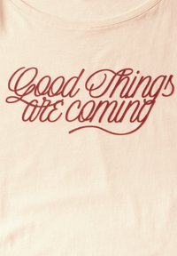 Camisa de algodón rosa claro con texto en cursiva roja que dice "Cosas Buenas Están Por Venir". Tejido suave con un clásico escote de cuello redondo.