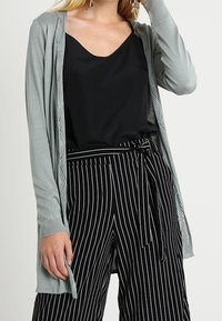 Světle šedý pletený cardigan s otevřeným předním dílem, černý saténový top s klesajícím krkem a černé pruhované kalhoty s vysokým pasem a uvázaným pasem.