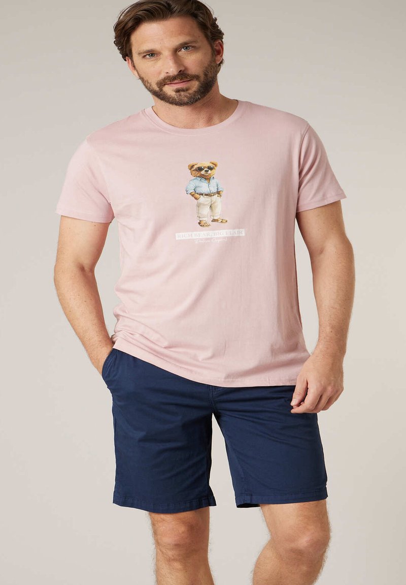 Homme portant un t-shirt rose clair avec un motif d'ours et un short bleu marine, debout avec une main dans la poche, sur un fond uni.