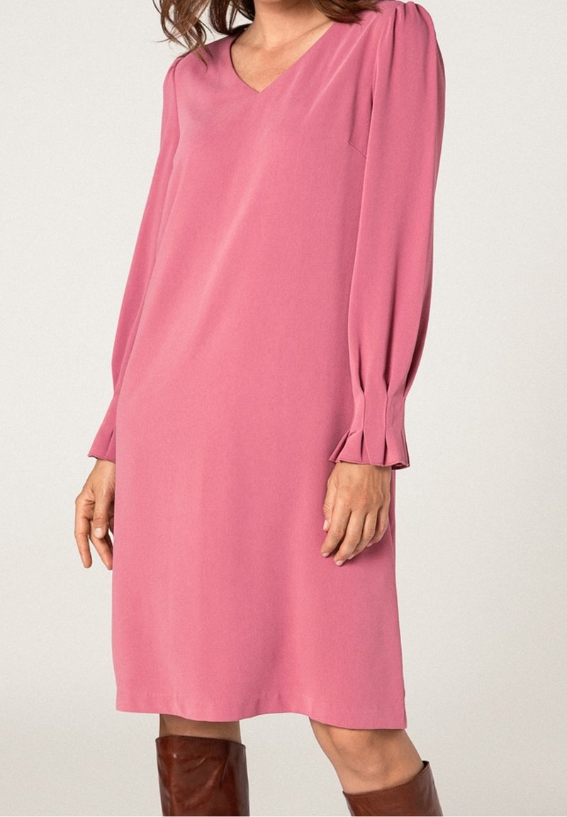 Femme portant une robe rose mi-longue à manches longues, col en V et poignets plissés, associée à des bottes marron montant jusqu’aux genoux.