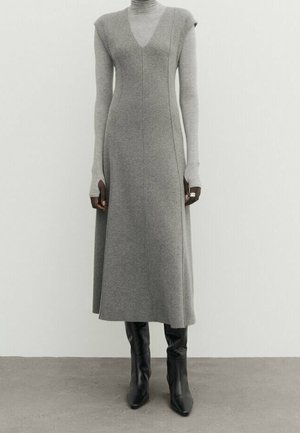 Robe midi grise sans manches à col en V portée sur un col roulé gris clair, associée à des bottes noires à talons montant jusqu'aux genoux, sur un fond uni.