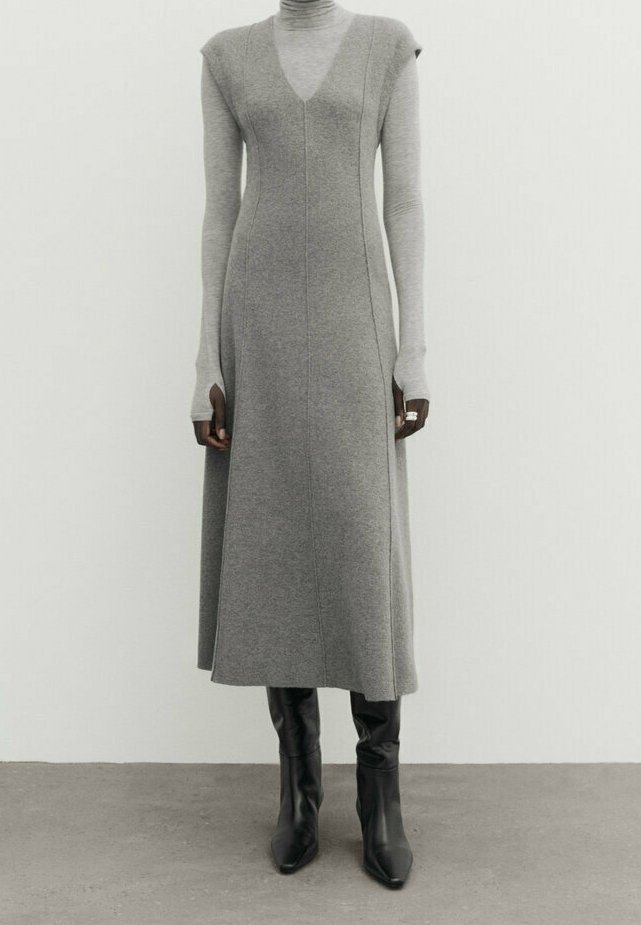 Robe midi grise sans manches à col en V portée sur un col roulé gris clair, associée à des bottes noires à talons montant jusqu'aux genoux, sur un fond uni.