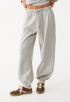 Persoon draagt losse lichtgrijze joggingbroek met trekkoord en elastische boorden, gecombineerd met bruine en crèmekleurige sneakers, staand tegen een witte achtergrond.