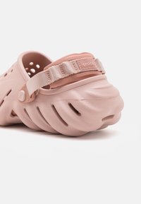 Light pink Crocs featuring a closed-toe design, ventilation holes, and a soft fabric heel strap with a logo patch. Textured material.