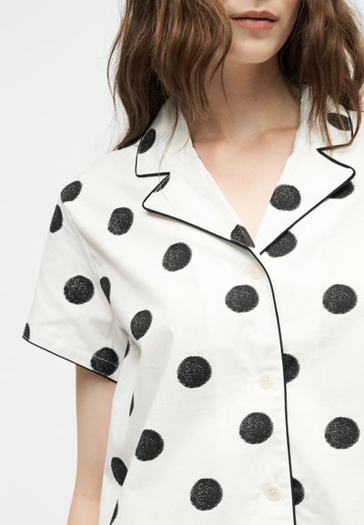 Femme portant une chemise blanche à manches courtes avec de gros pois noirs et un liseré noir le long du col et de la couture des boutons.