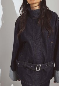 Woman wearing dark denim jacket with high neck, belt with silver buckle, and turned-up gray cuffs, standing against a plain background.