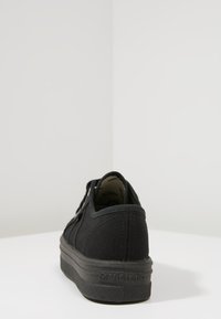 Sneaker nere a basso profilo con suola spessa e logo "Victoria" ben visibile sul tallone, posizionata su una superficie bianca contro un muro angolare semplice.