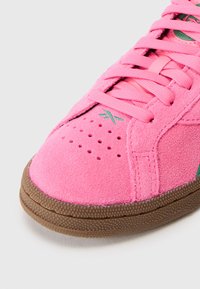 Αθλητικά παπούτσια με ροζ suede επάνω μέρος, στρογγυλή μύτη, περίπλοκες τρύπες στο ύφασμα, ελαφρώς ανυψωμένη σόλα από καουτσούκ, και πράσινες λεπτομέρειες στο πλάι.