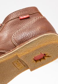 Chaussure en cuir brun avec une finition texturée, semelle en caoutchouc jaune avec un design découpé, et une étiquette rouge "Kickers" sur le côté.