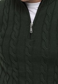 Ciemnozielony dziergany sweter z kołnierzem na półzamek, z wzorem warkoczowym i teksturowanym wykończeniem. Srebrny suwak.