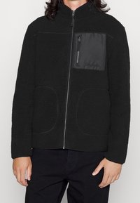 Veste en polaire noire avec une fermeture éclair à l'avant, un col montant et une poche plaquée avec un tissu lisse. Deux poches frontales arrondies.