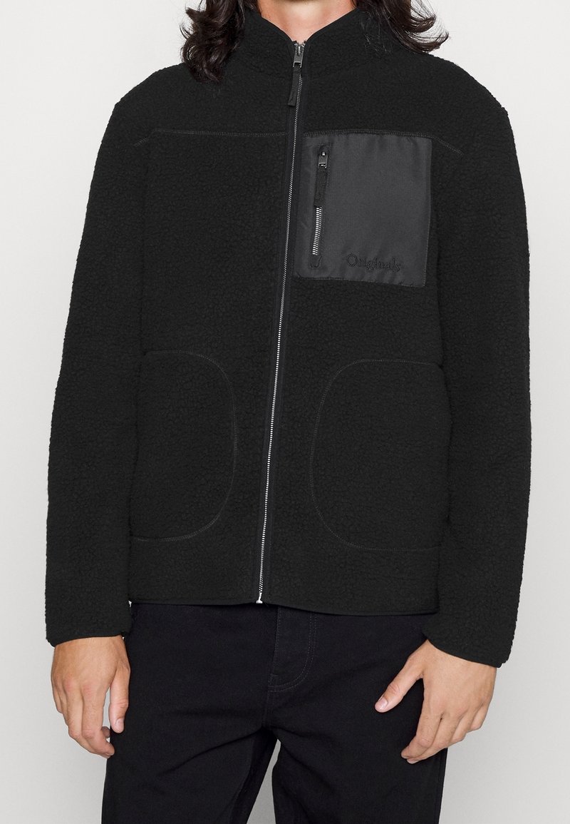 Veste en polaire noire avec une fermeture éclair à l'avant, un col montant et une poche plaquée avec un tissu lisse. Deux poches frontales arrondies.