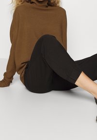 Brązowy sweter z golfem, zestawiony z czarnymi spodniami typu cropped. Miękka tekstura, luźny krój, widoczne podwinięcia na nogawkach.