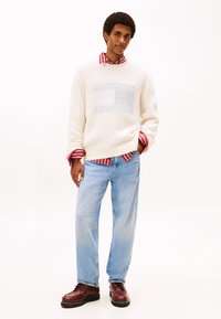 Άσπρο πλεκτό πουλόβερ με υφή λογότυπου, φορεμένο πάνω από μια κόκκινη και άσπρη ριγέ πουκάμισο. Φόδρα ανοιχτό μπλε τζιν και σκούρα μπορντό παπούτσια συμπληρώνουν το ντύσιμο.