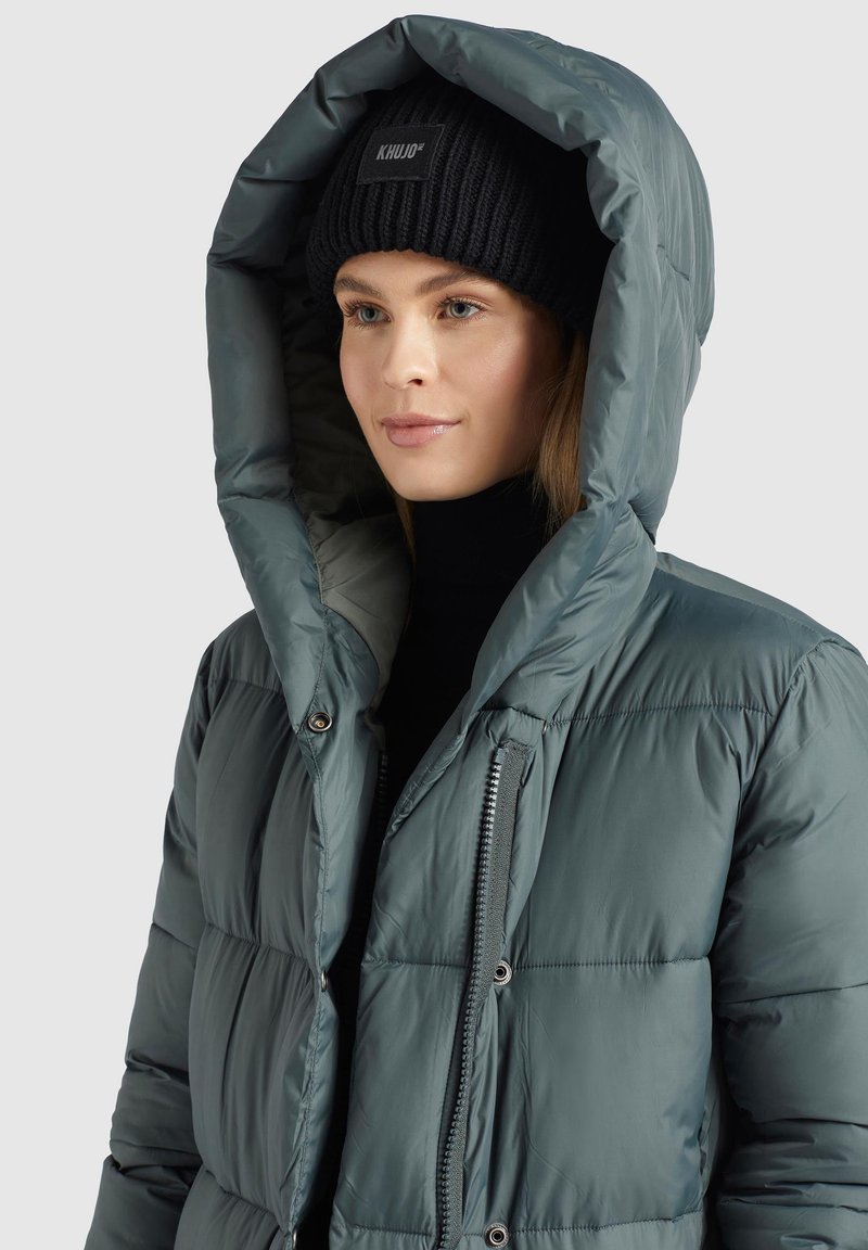 Dunkelgrüne Pufferjacke mit großer Kapuze, Reißverschluss und elastischen Bündchen; getragen mit einer schwarzen Strickmütze. Glatte, glänzende Textur.