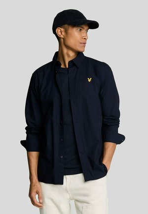 Man draagt een zwarte pet, een marineblauw overhemd met gele logo en knopen, een zwart T-shirt en een witte broek, kijkt opzij met één hand in de zak.