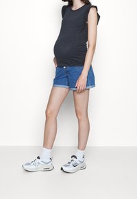 Těhotenské tričko v tmavě šedé barvě, bez rukávů s volánkovými detaily, kombinované s ohrnutými denimovými šortkami a bílými teniskami se šedými akcenty.