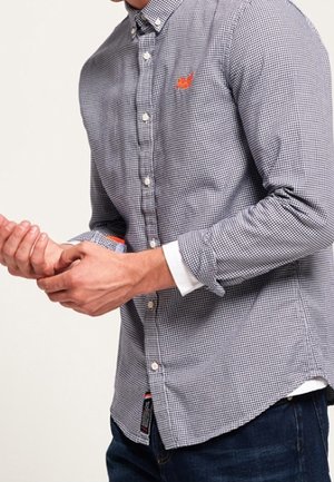 Mann trägt ein kariertes Hemd mit Knöpfen und hochgekrempelten Ärmeln, richtet ein Handgelenk, sichtbar vom Hals bis zur Taille.