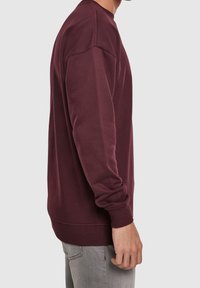 Uomo che indossa una felpa a maniche lunghe color bordeaux e jeans grigi chiari, mostrato di lato su uno sfondo grigio uniforme.