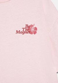 Svetlo roza tkanina z rdečim napisom »Bodi magnet« in grafiko rdečega hibiskusa na zgornjem delu prsnega koša.