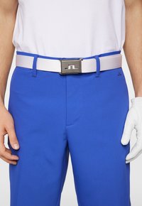 Pantaloni blu reale con una cintura bianca dotata di una fibbia metallica argento. Un guanto da golf è indossato sulla mano destra, enfatizzando il design sportivo.
