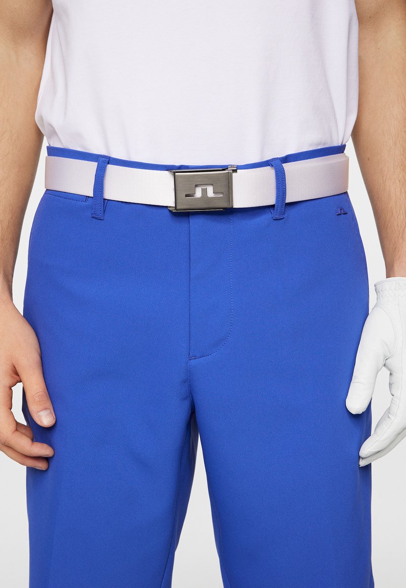 Pantaloni blu reale con una cintura bianca dotata di una fibbia metallica argento. Un guanto da golf è indossato sulla mano destra, enfatizzando il design sportivo.