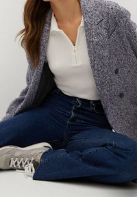 Donna seduta con le gambe incrociate, indossa un blazer grigio testurizzato, una maglia bianca con zip, jeans blu scuro e sneakers chiare.