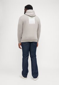 Szary kaptur z dużym białym logo Calvin Klein na plecach, zestawiony z ciemnoniebieskimi dżinsami i białymi trampkami. Gładki materiał i luźny krój.