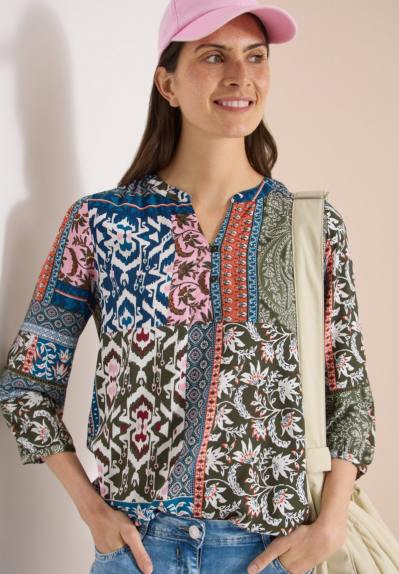 Femme portant une casquette rose et un chemisier coloré à motifs floraux et géométriques, un jean bleu et un sac à bandoulière beige clair.