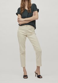 Béžové džíny s vysokým pasem a zúženými nohavicemi, s knoflíkovým a zipovým zapínáním, spárované s volným tmavozeleným tričkem a černými sandály na podpatku.