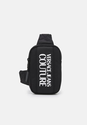 Sac bandoulière noir Versace Jeans Couture avec texte de logo vertical blanc et poche frontale zippée sur fond uni.