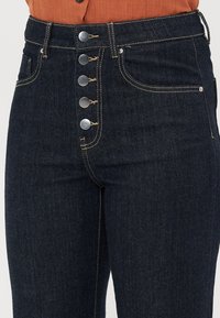 Mørke denim høyt liv jeans med fem-knapp lukking, kontrasterende gyllen søm, og frontlommer. Glatt tekstur av stoffet.