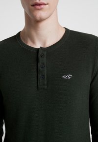 Camisa de manga larga de tejido waffle en color verde, con cuello redondo, cuatro botones negros y un pequeño logotipo blanco en el lado izquierdo del pecho.