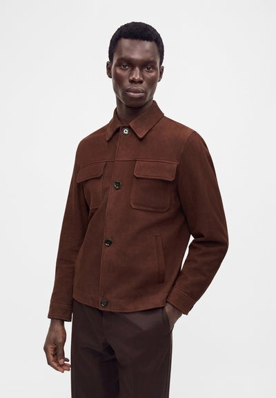 Homme portant une veste boutonnée marron foncé avec poches à rabat et un pantalon marron foncé assorti, debout devant un fond clair uni.