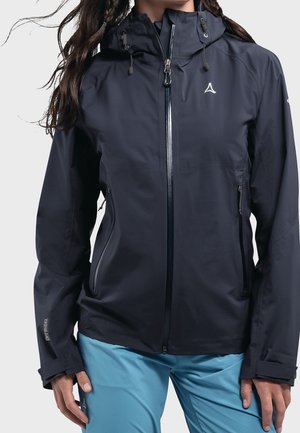 Vrouw draagt een zwarte waterdichte jas met capuchon en ritssluiting aan de voorkant en een blauwe broek, staand tegen een effen achtergrond.