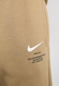 Brune bomull joggebukser med en hvit Nike swoosh-logo og svart tekst med selskapets adresse trykt på siden.