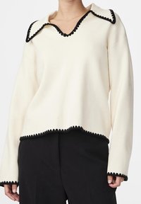 Cremiger Pullover mit langen Ärmeln und tiefem V-Ausschnitt, verziert mit einem dekorativen schwarzen Wellenrand am Kragen und Saum für zusätzliche Details.