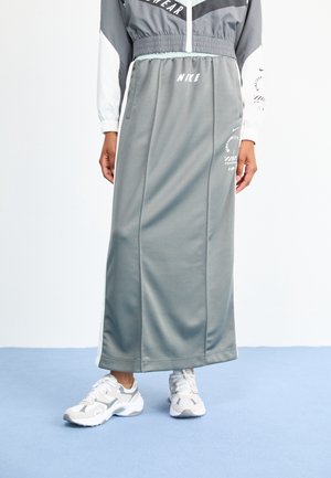 Femme portant une jupe de sport grise Nike et une veste assortie avec des chaussures de sport blanches et beiges, debout sur un sol bleu clair.