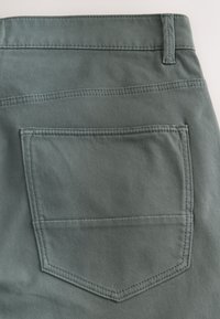 Poche arrière et passants de ceinture sur un pantalon en denim vert délavé avec des coutures visibles et un tissu texturé.