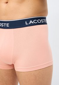 Κοντινό πλάνο ενός ατόμου που φορεί ροζ ανδρικά εσώρουχα τύπου boxer με μπλε ναυτικό μέση που φέρει το λευκό λογότυπο της Lacoste.