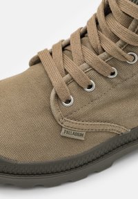 Zapatilla alta de lona marrón con superficie texturizada, ojales de metal y cordones planos. Cuenta con suela de goma y una pequeña etiqueta "PALLADIUM".