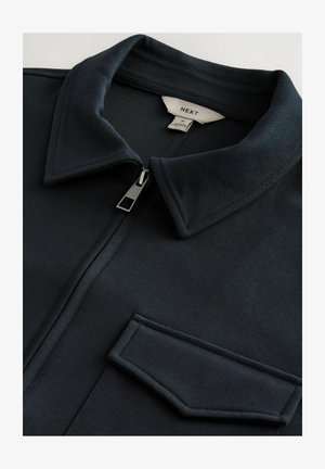 Granatowa kurtka zapinana na zamek, z ostrym kołnierzem, teksturowanym materiałem i jedną kieszenią na piersi. Posiada metalowy zamek błyskawiczny oraz mocne szwy.