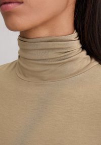 Nahaufnahme des Nackens und der Lippen einer Person, die ein beiges, geripptes Rollkragenshirt trägt, mit langen dunklen Haaren, die sichtbar sind.