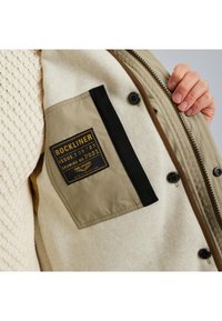 Béžový a krémový interiér bundy s čiernou kapsou obsahujúcou zlatú a čiernu nášivku; obsahuje gombíky a textúrovanú podšívku.