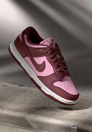 Niski model butów Nike w różowym i bordowym skórzanym wykończeniu z brązowymi sznurowadłami, logo Swoosh oraz białą podeszwą, prezentowany na betonowej powierzchni.